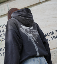 Vintage washed black streetwear hoodie with angel figure back graphic and minimalist logo design shown in an outdoor lifestyle setting.