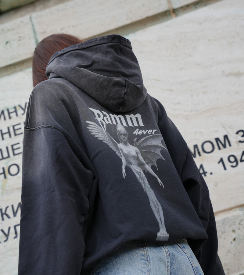 Vintage washed black streetwear hoodie with angel figure back graphic and minimalist logo design shown in an outdoor lifestyle setting.