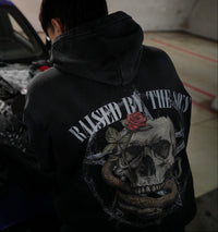 Vintage washed black streetwear hoodie with raised by the 80s back graphic featuring skull, snake and rose design shown in a lifestyle setting.