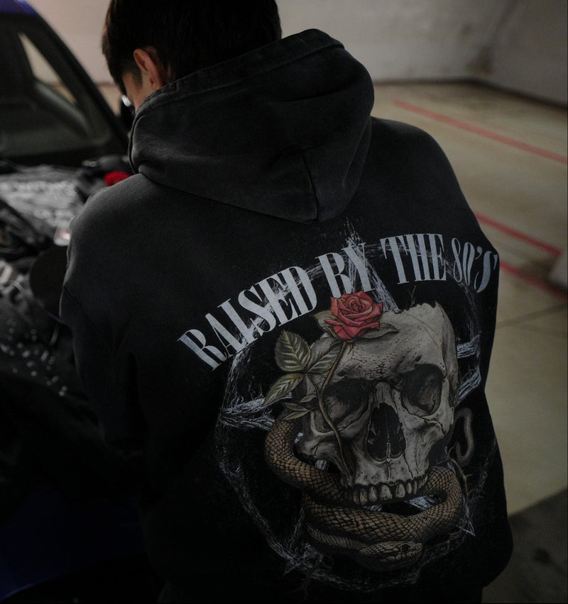 Vintage washed black streetwear hoodie with raised by the 80s back graphic featuring skull, snake and rose design shown in a lifestyle setting.